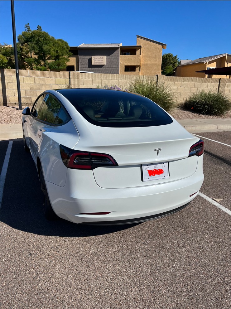 The 2023 Tesla Model 3 Standard Range Plus RWD sitting slightly elevated in a parking lot. Its doors and hood are closed. The car has a signature Tesla roof rack.